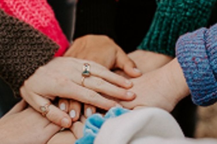 Viele Hände liegen Aufeinander als Zeichen des Zusammenhalts. Die Hand ganz oben trägt einen Ring am Ringfinger. 